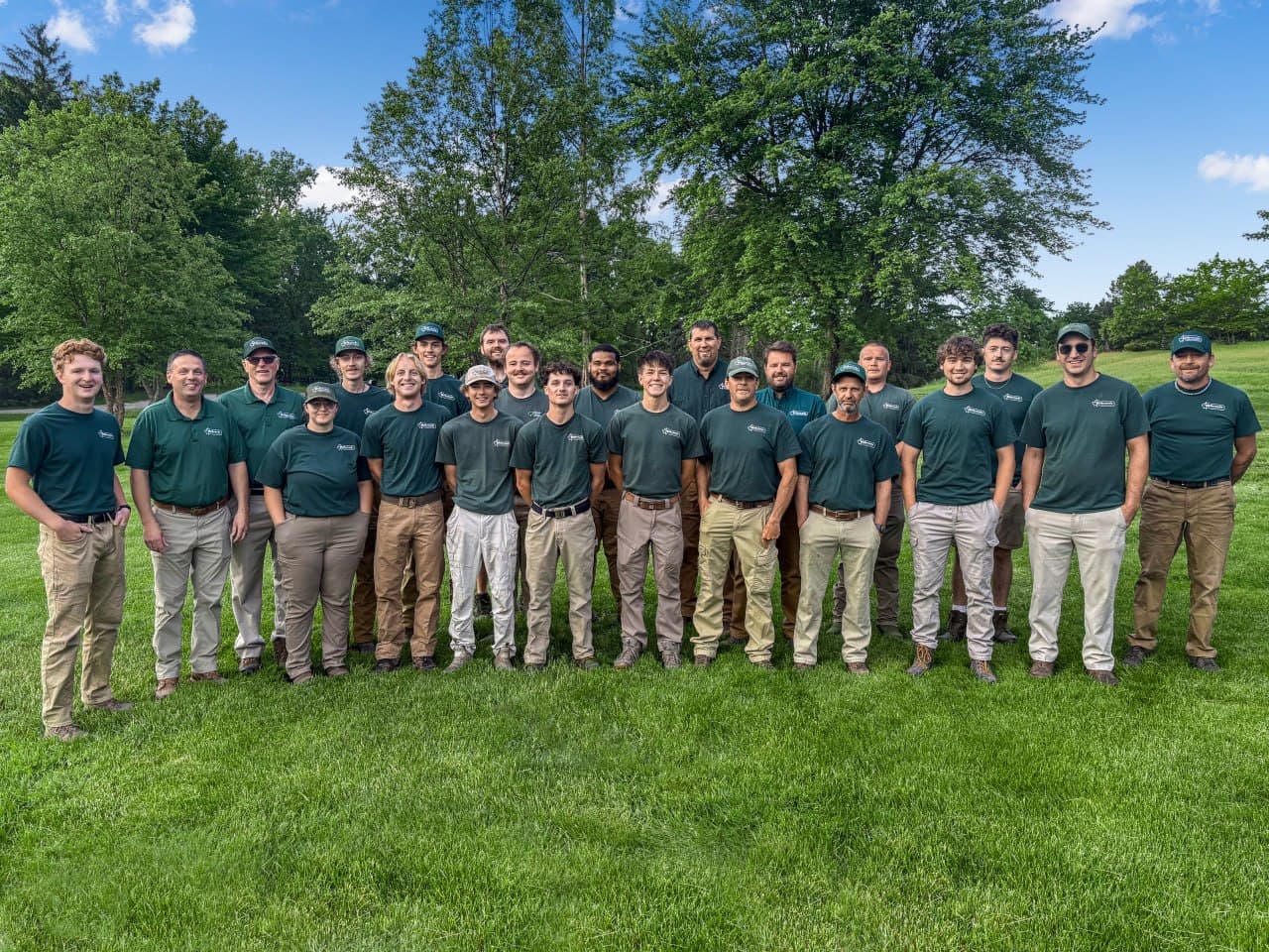 The Hilbrands crew — eighteen team members in green Hilbrands shirts standing together on a kept lawn in front of mature trees under a bright blue sky.