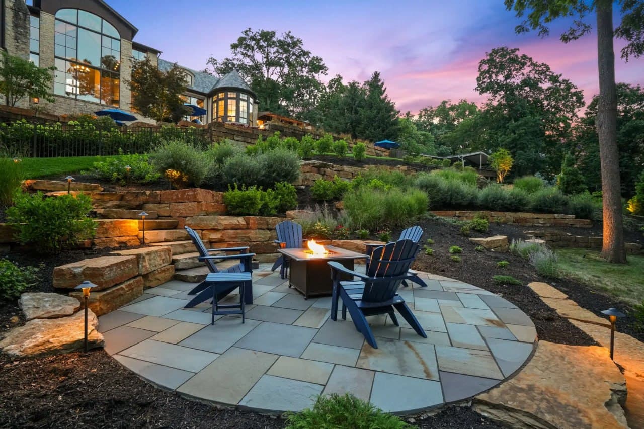 Landscape enhancement — stacked-stone tiered retaining walls with path lighting, mature plantings, and a circular bluestone patio with fire table and Adirondack seating in front of an estate at twilight