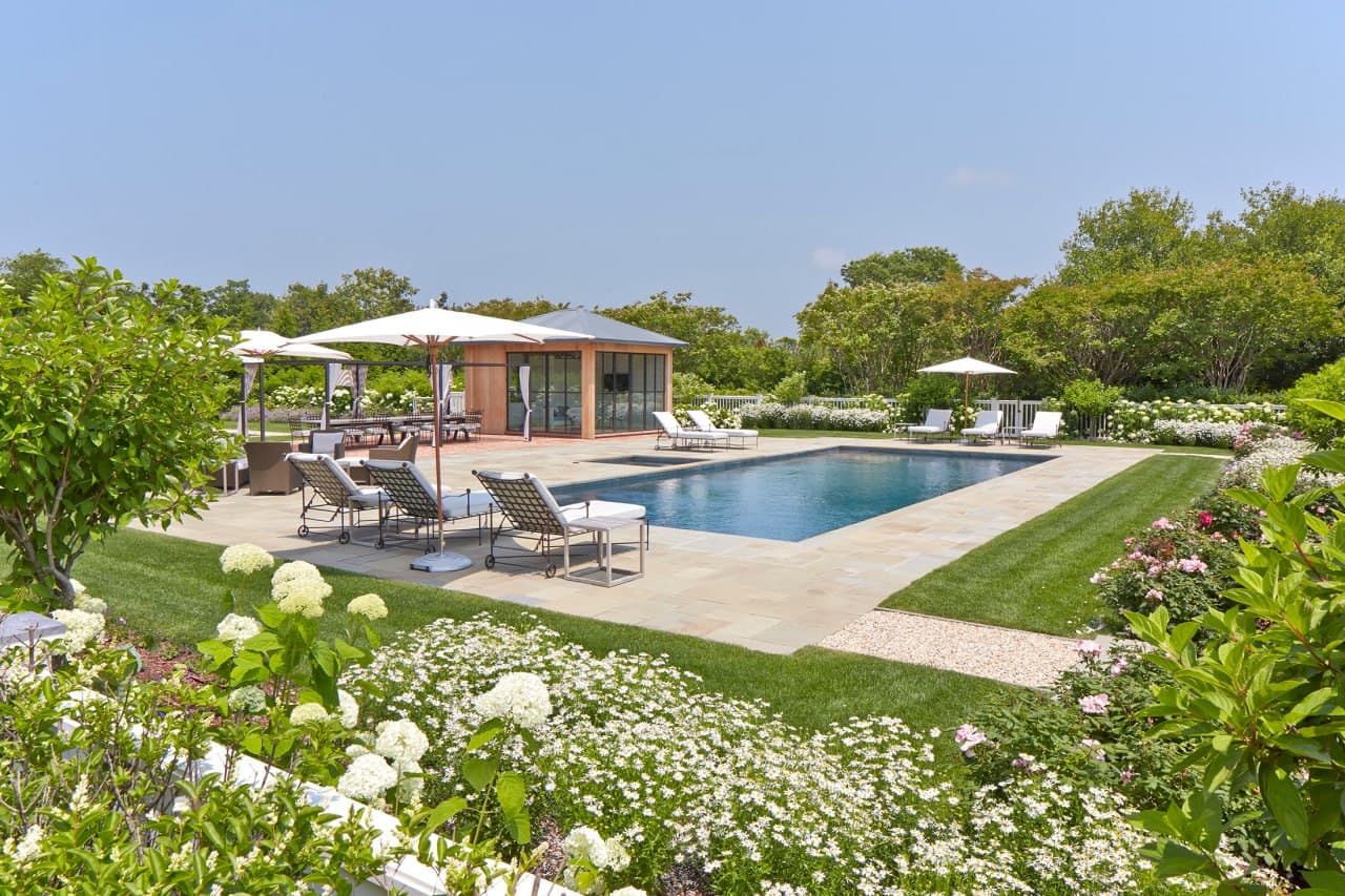 Landscape enhancement detail — rectangular pool with bluestone deck and modern wood-and-glass cabana, framed by mass hydrangea hedges and mixed plantings on a sunny day