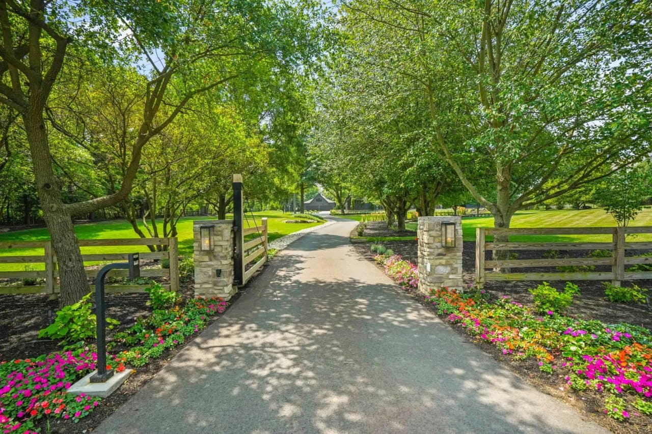 Tree-canopied driveway gate flanked by stone columns and impatiens borders.