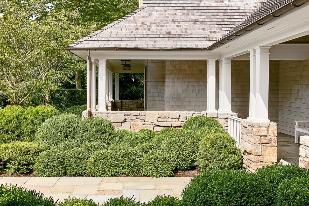 Cedar-shingle porch with stone footing and rounded boxwood plantings.