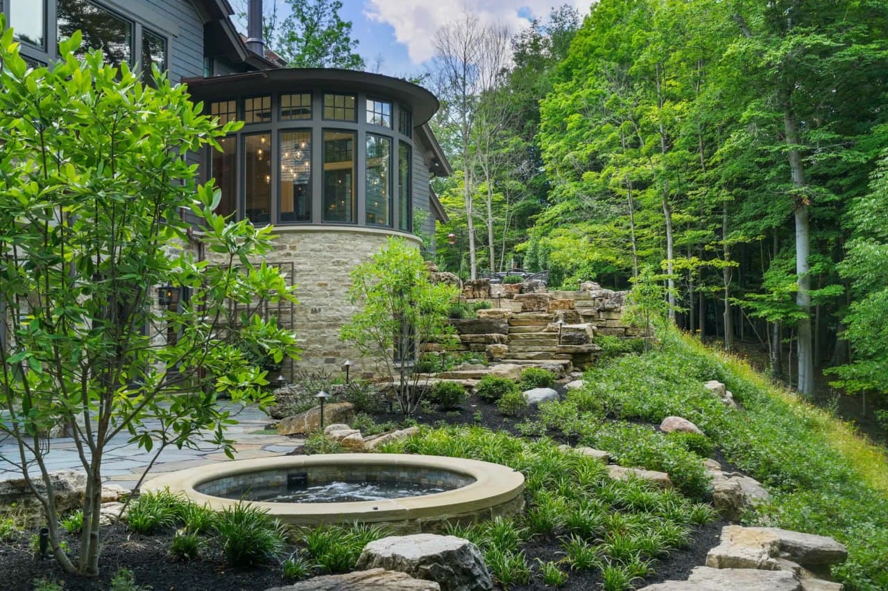 Bluestone slab steps cutting up a hillside through fresh garden beds toward a shingled cape with stone chimney.