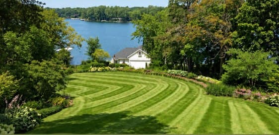 Curved striped lawn descending toward a lakeside cottage with mature trees and waterside framing.